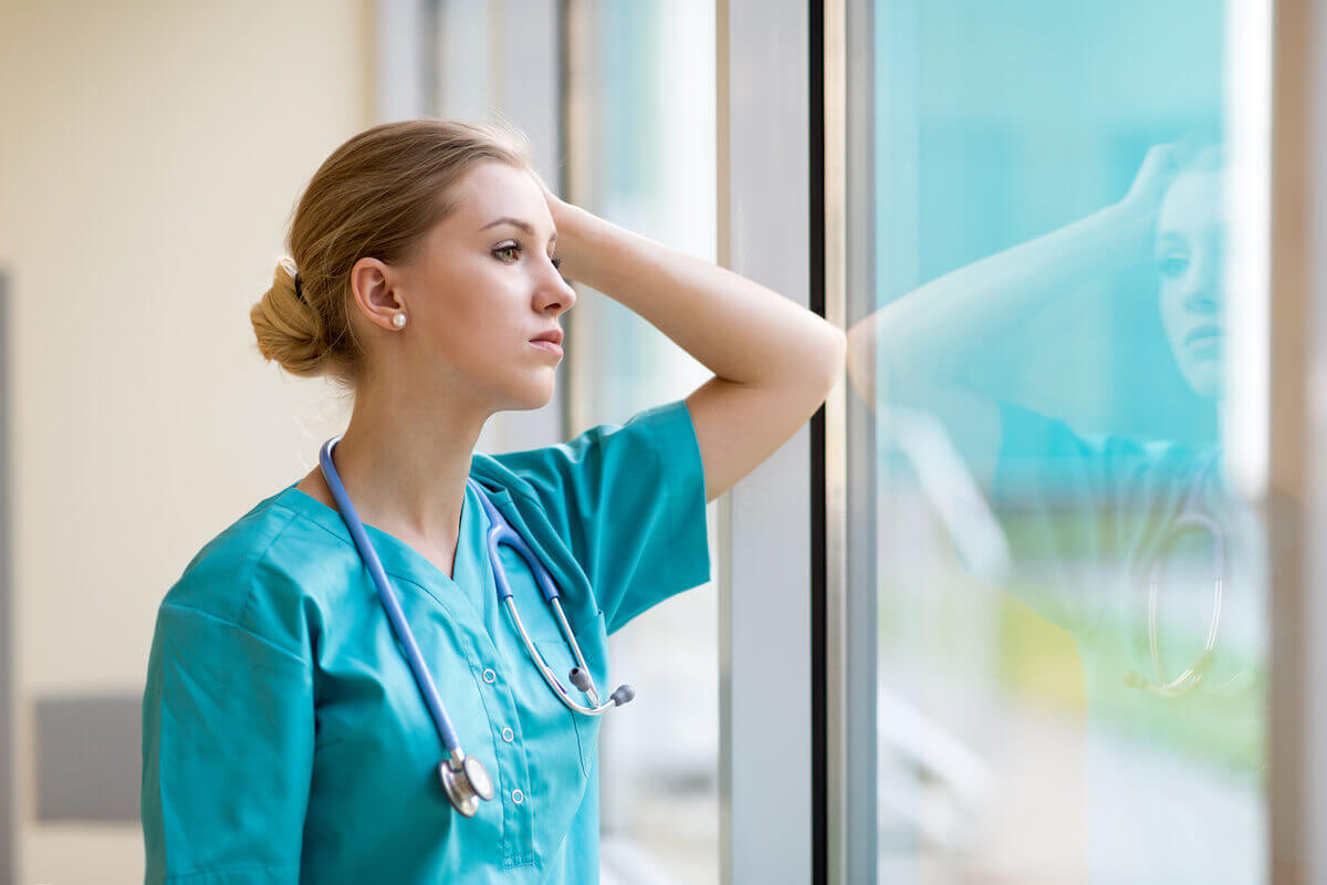 Young nurse with serious expression leaning elbow against a window and looking outside.