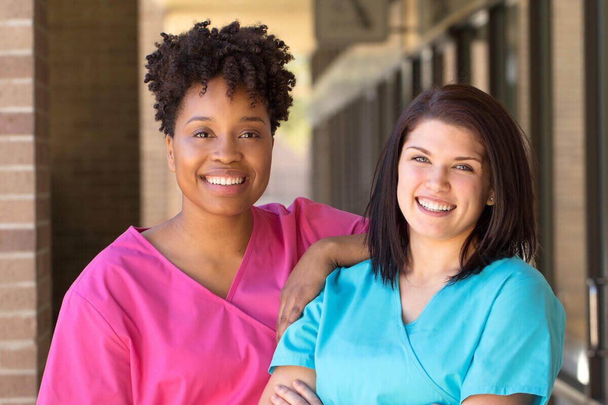 African-American and white nurse smiling and standing together outside a facility.