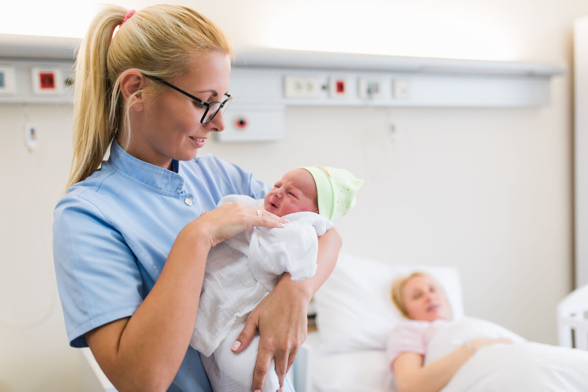 Female nurse in glasses holding newborn with recovering mother in background.