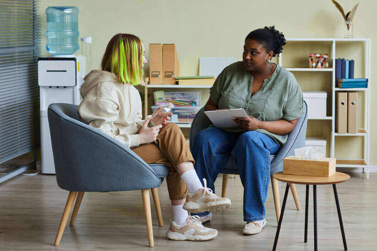 African-American social worker sitting with student who has bright green hair in a classroom.