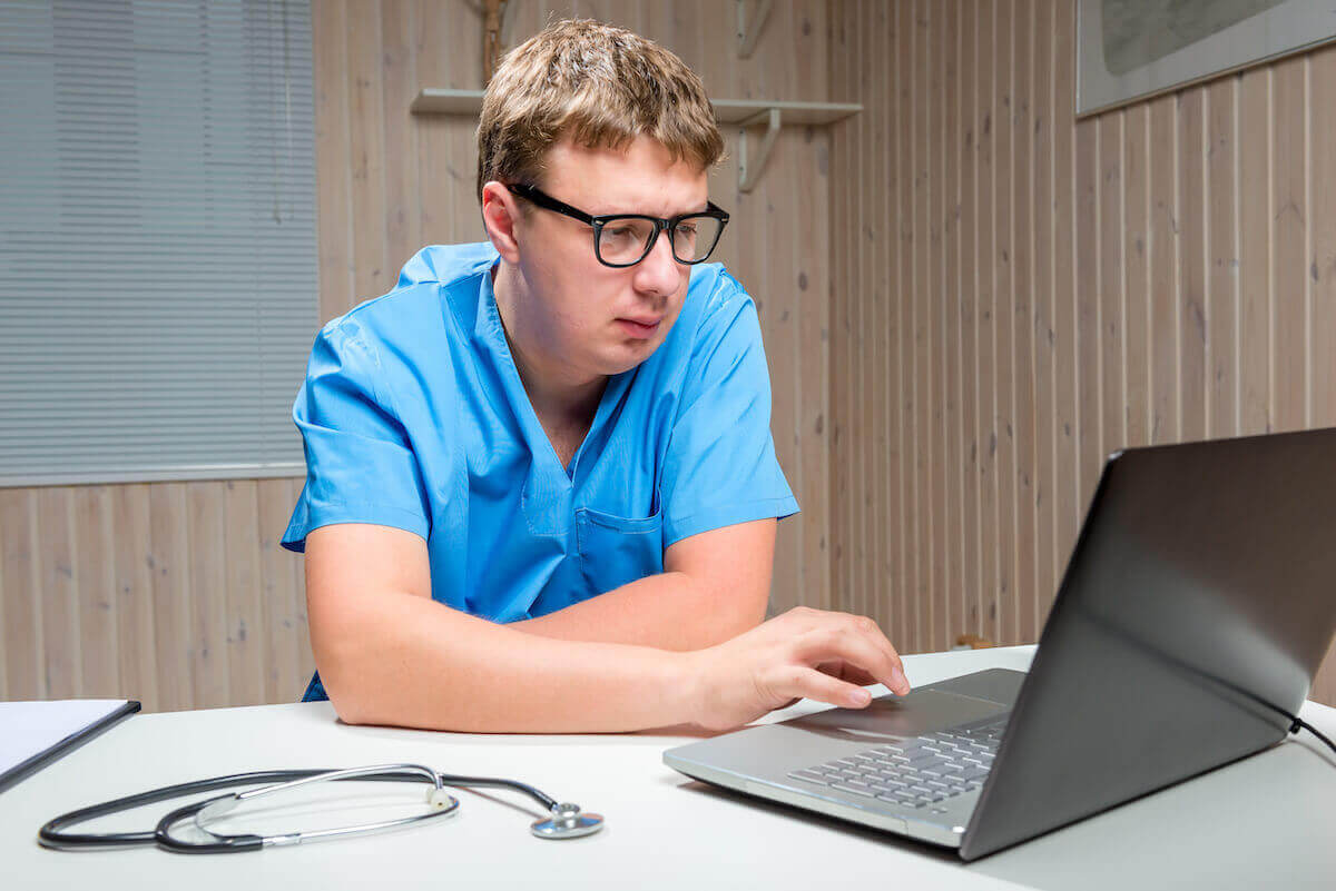 Male nurse with glasses and blue scrubs working on a laptop in home office.