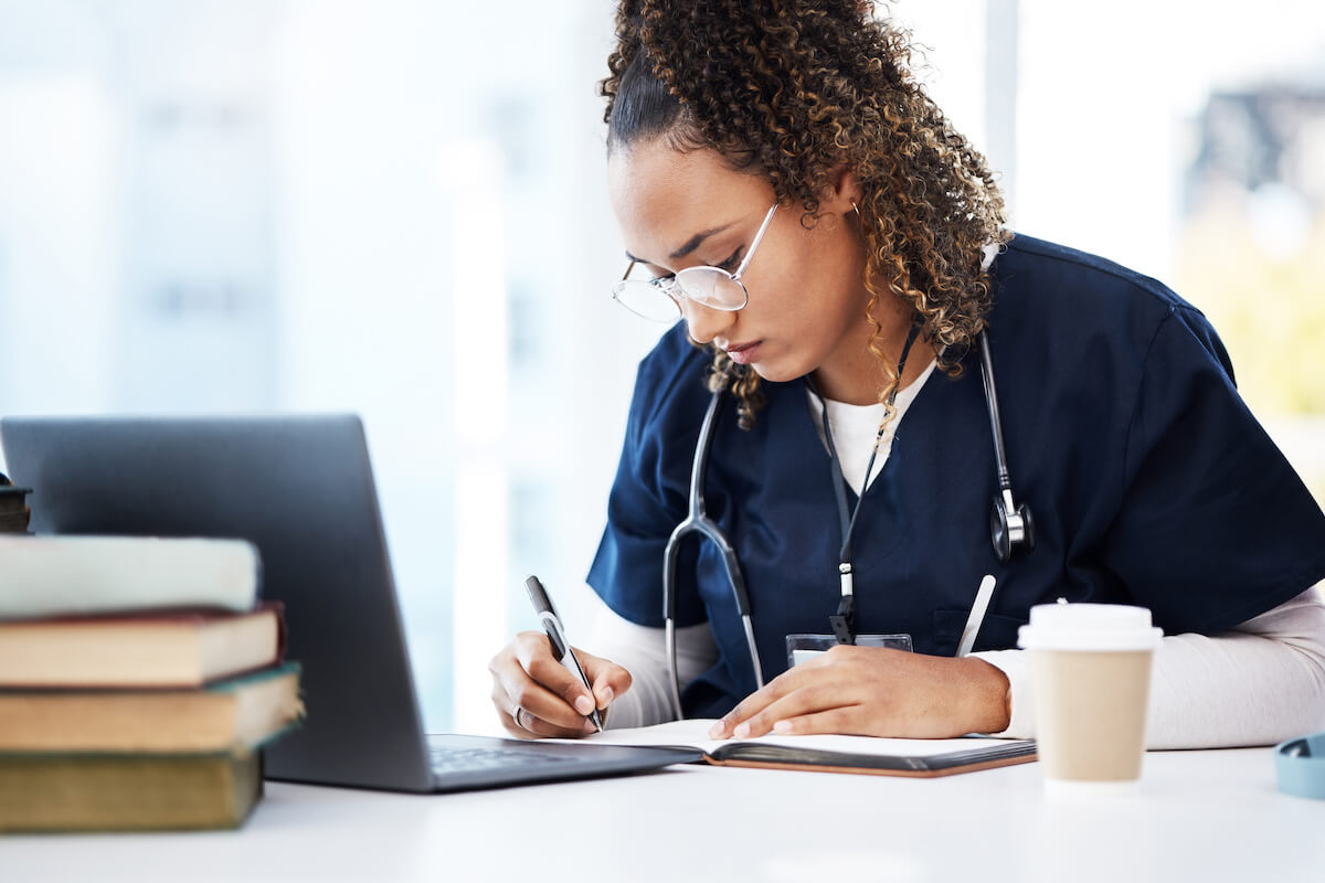 Young nurse writing in a journal in front of a laptop with a cup of coffee.