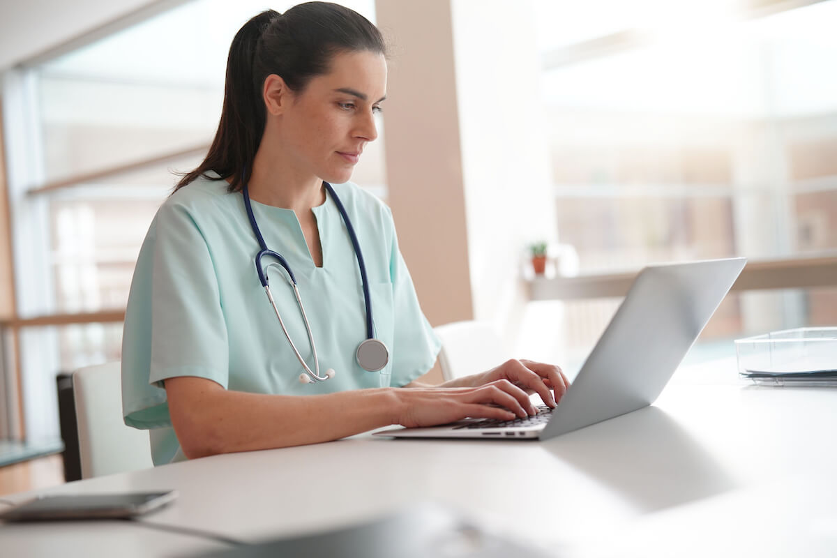 Young nurse with hair tied back sitting at desk and working on laptop.