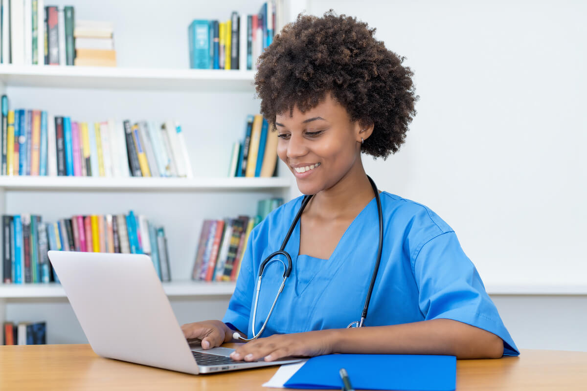 Nurse smiling and sitting at laptop with bookshelf behind her.
