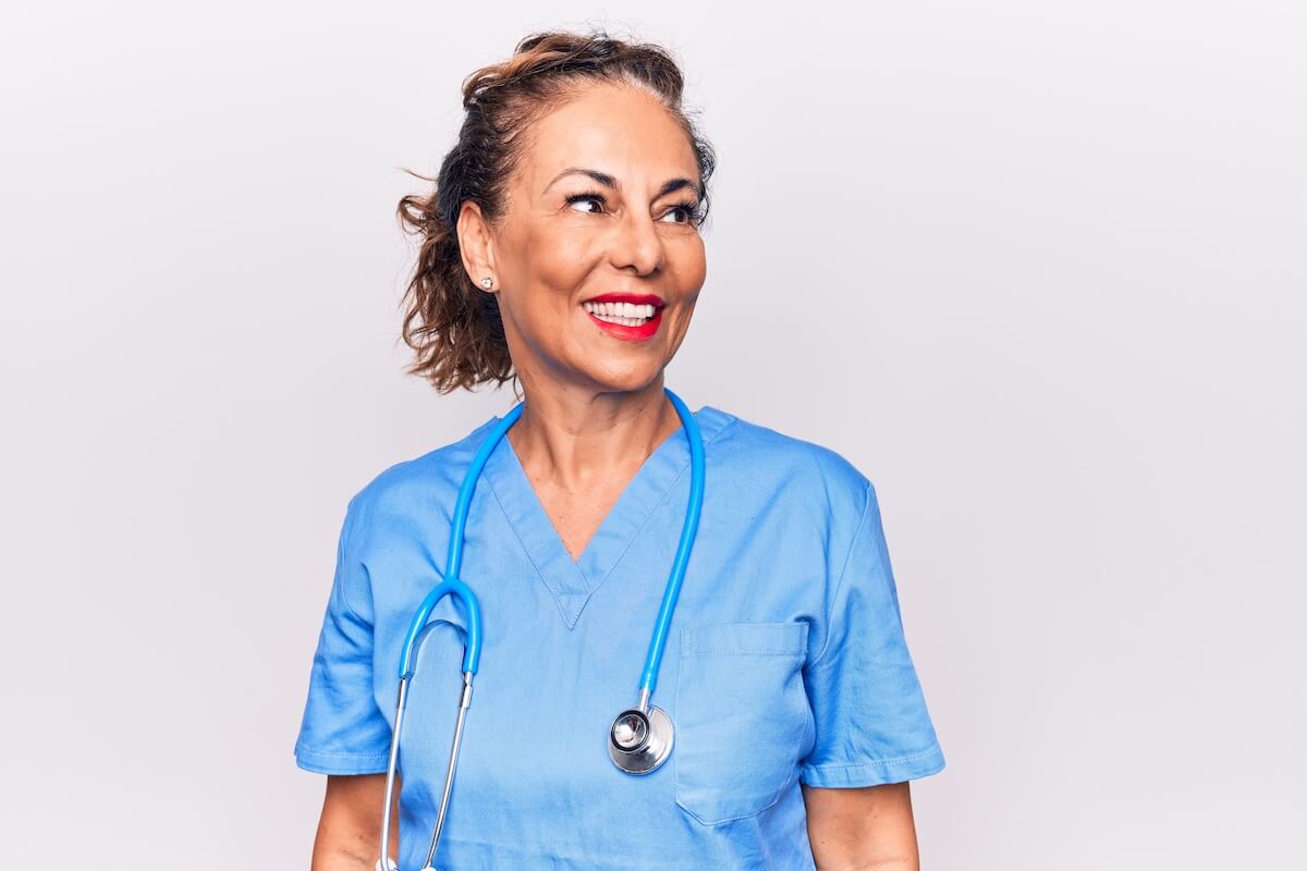 Older female nurse with blue scrubs looking to the side and smiling.