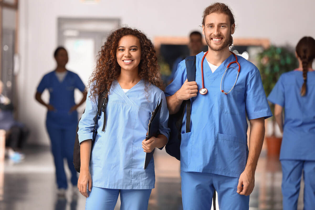 Young female and young male nurse in scrubs wearing backpacks and smiling.