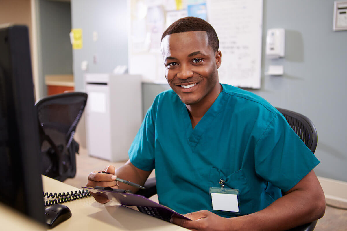 Male nurse with blue scrubs sitting behind a computer at work and smiling.