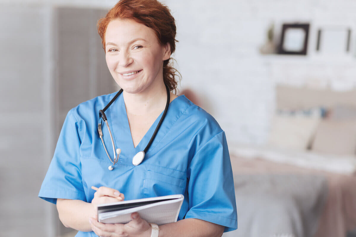Nurse with red hair and light blue scrubs smiling and holding a clipboard.