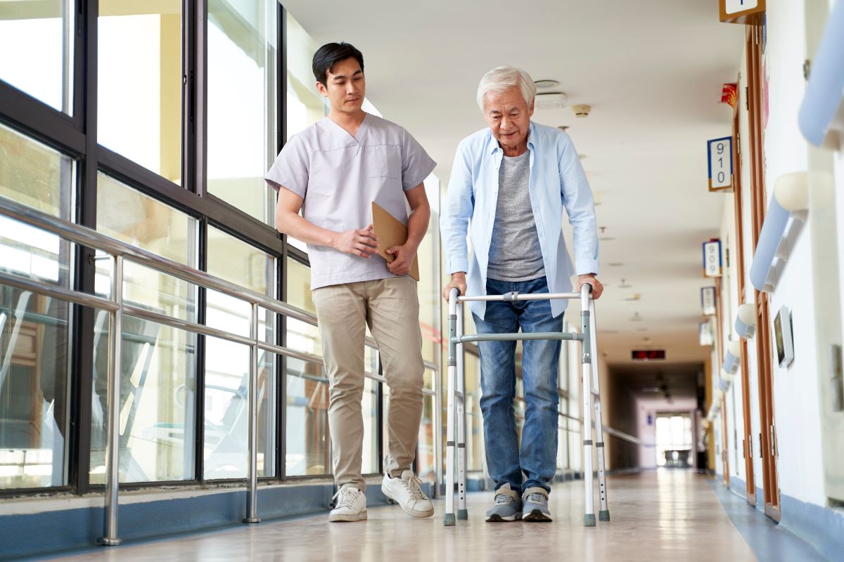 A nurse at a skilled nursing facility, or SNF, assists an elderly man using a walker.