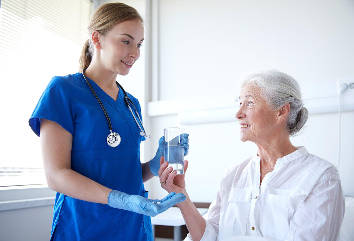 CMA nurse in blue scrubs handing pills and water to a senior patient.