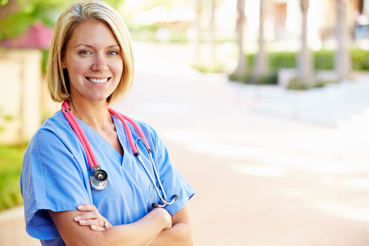 RN with short blonde hair outside with her arms crossed and waiting to complete continuing education requirements for nurses.