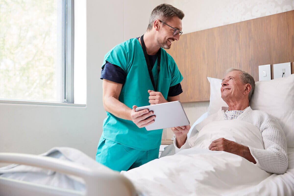 Male nurse in green scrubs showing a tablet to an elder man in bed.