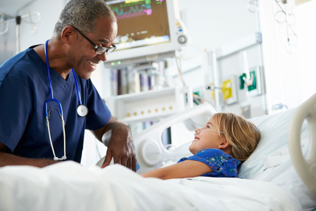 A nurse meets with a young girl lying in a hospital bed, and they share a smile.