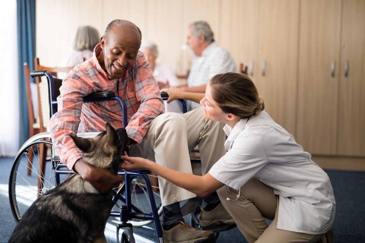 A nursing home resident, in a wheelchair, pets a dog while a nurse looks on.