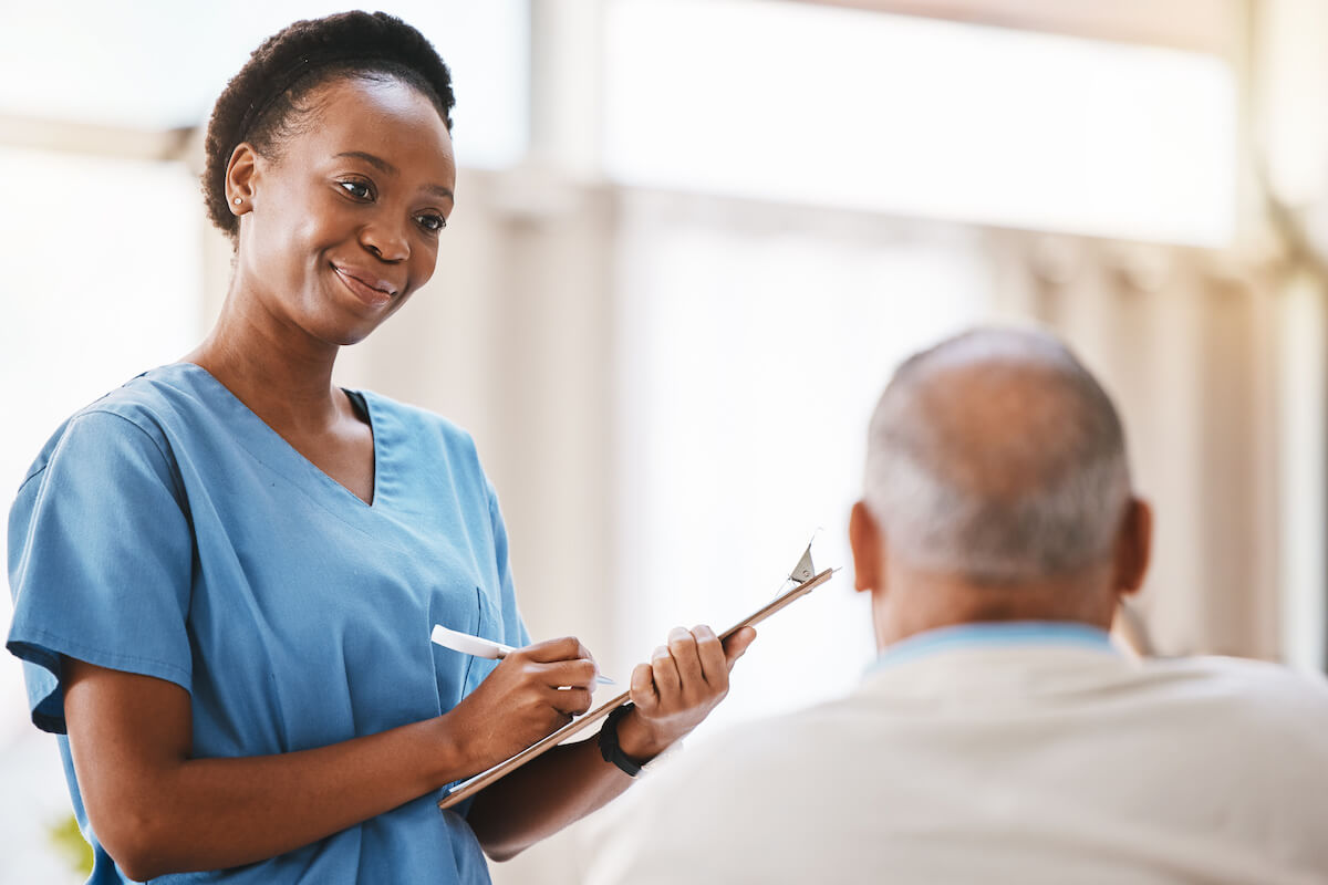 An African-American STNA with clipboard talking to a senior patient.