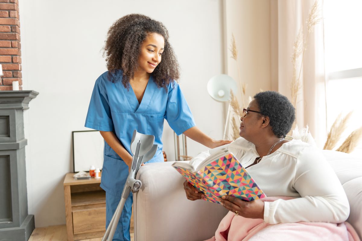 A nurse checks on a nursing home resident, who's sitting on a couch and reading a book.