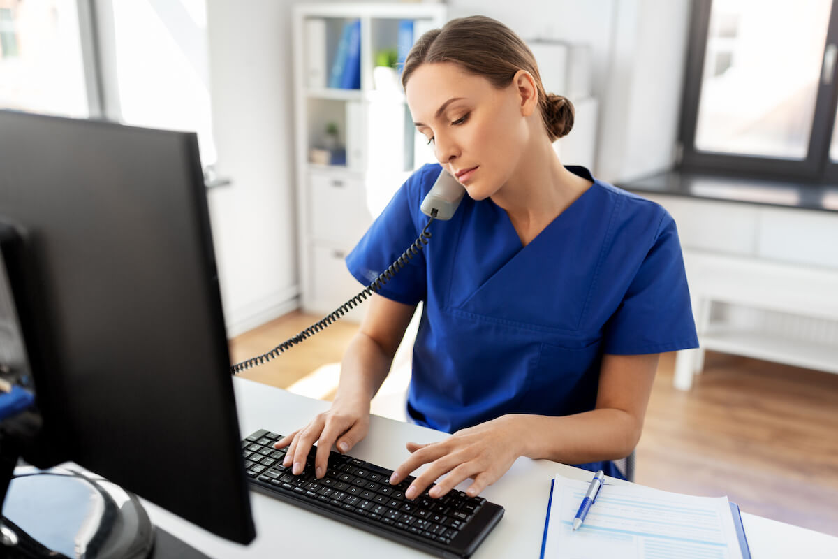 Nurse scheduler typing on a keyboard and holding a phone under her chin.