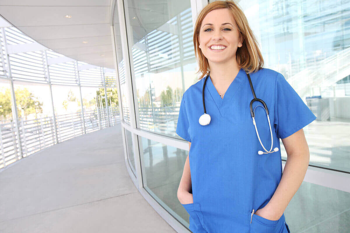 Travel nurse in blue scrubs standing outside of a hospital and smiling.