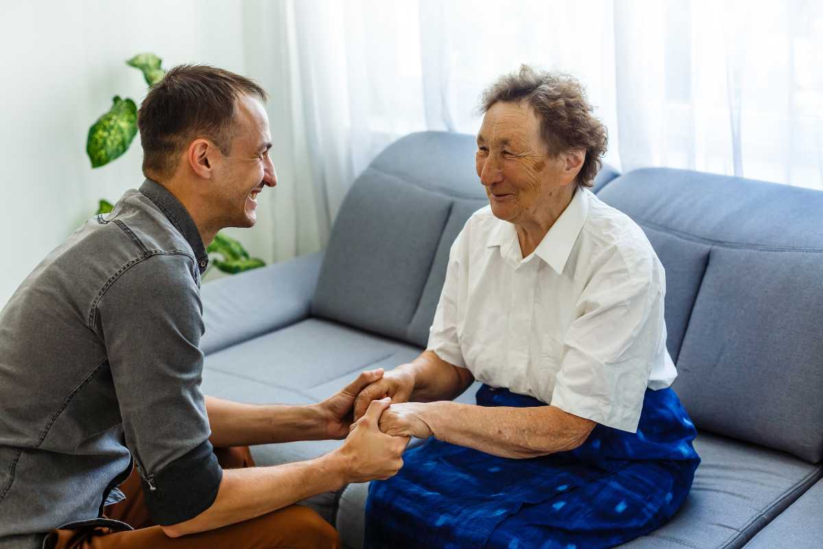 Caregiver holding hands with senior patient seated on a couch.