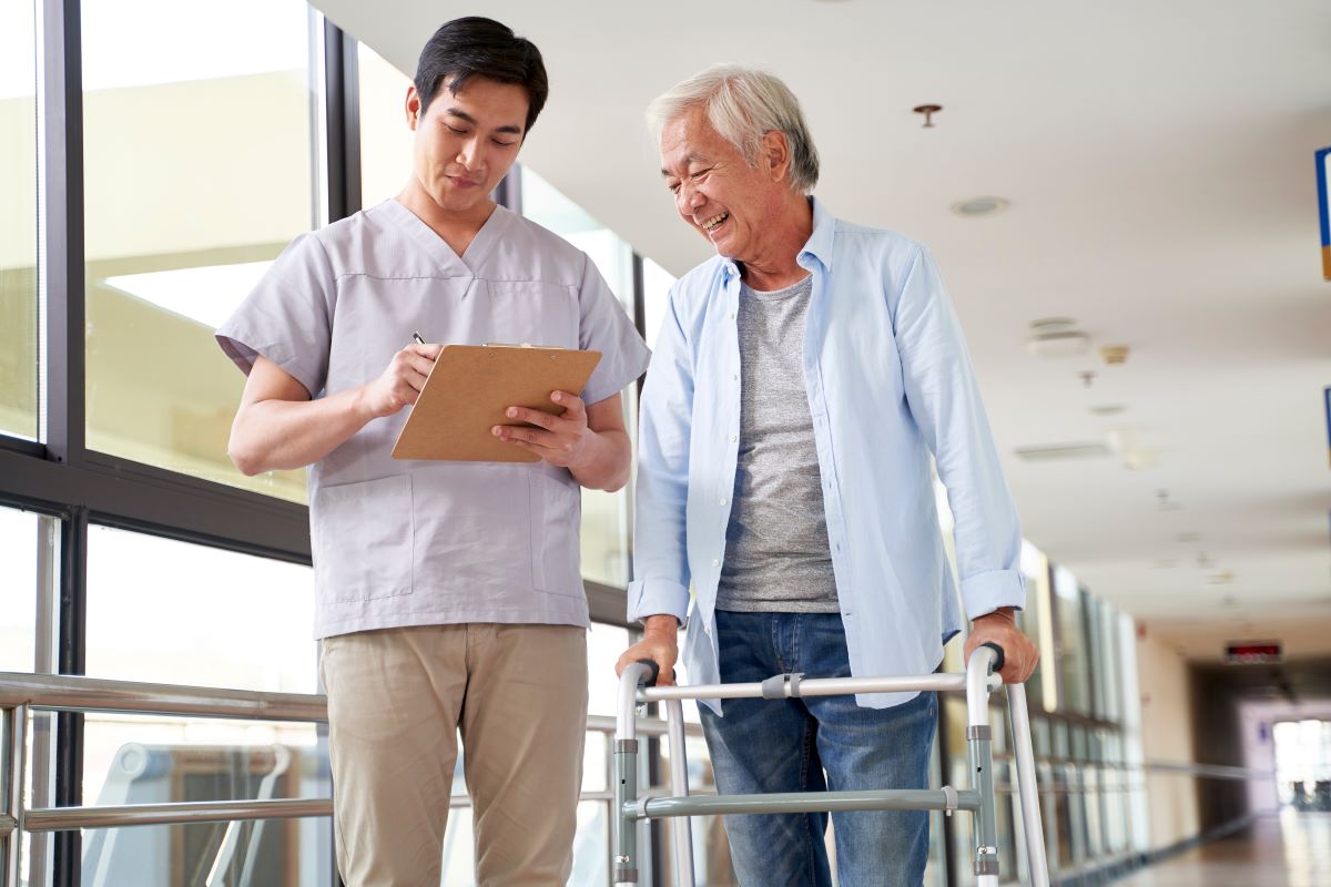 A nurse at a nursing home checks a resident's chart while standing with him in the hallway.