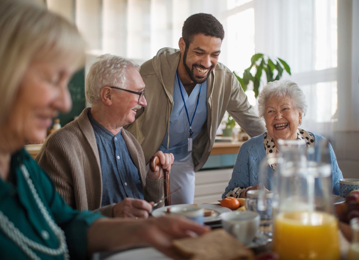 A nurse stands beside a group of nursing home residents, who are laughing as they share a meal together.