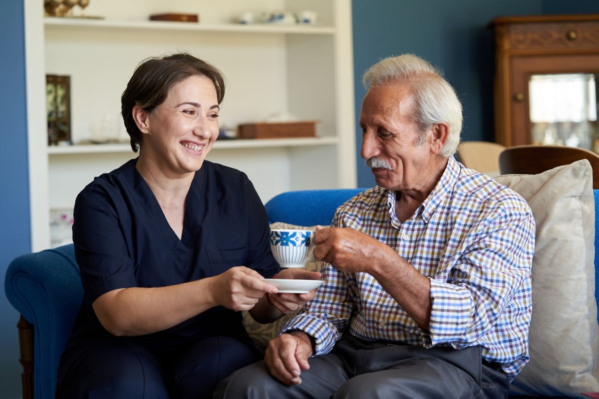 A caregiver at a nursing home helps a resident drink his tea.