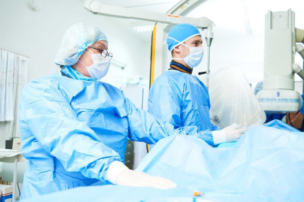 Cath lab nurses in operating room wearing scrubs, masks, gloves, and surgical gowns.