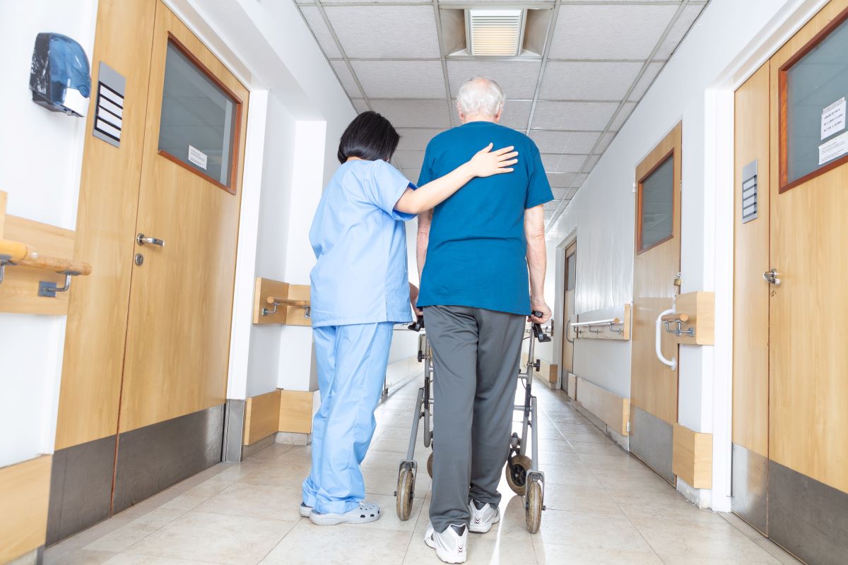 A nursing home care worker helps a resident walk down the hallway.