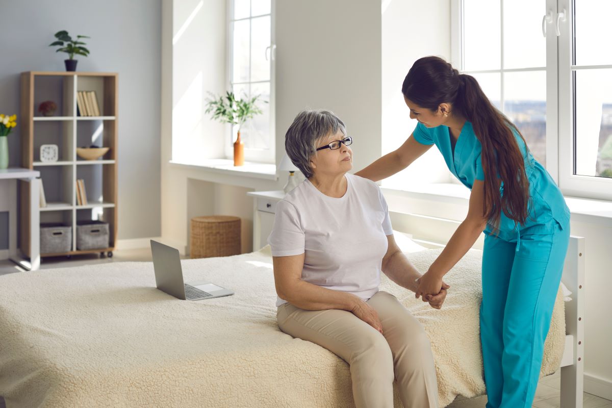 A nurse speaks with a nursing home resident who's sitting on her bed.