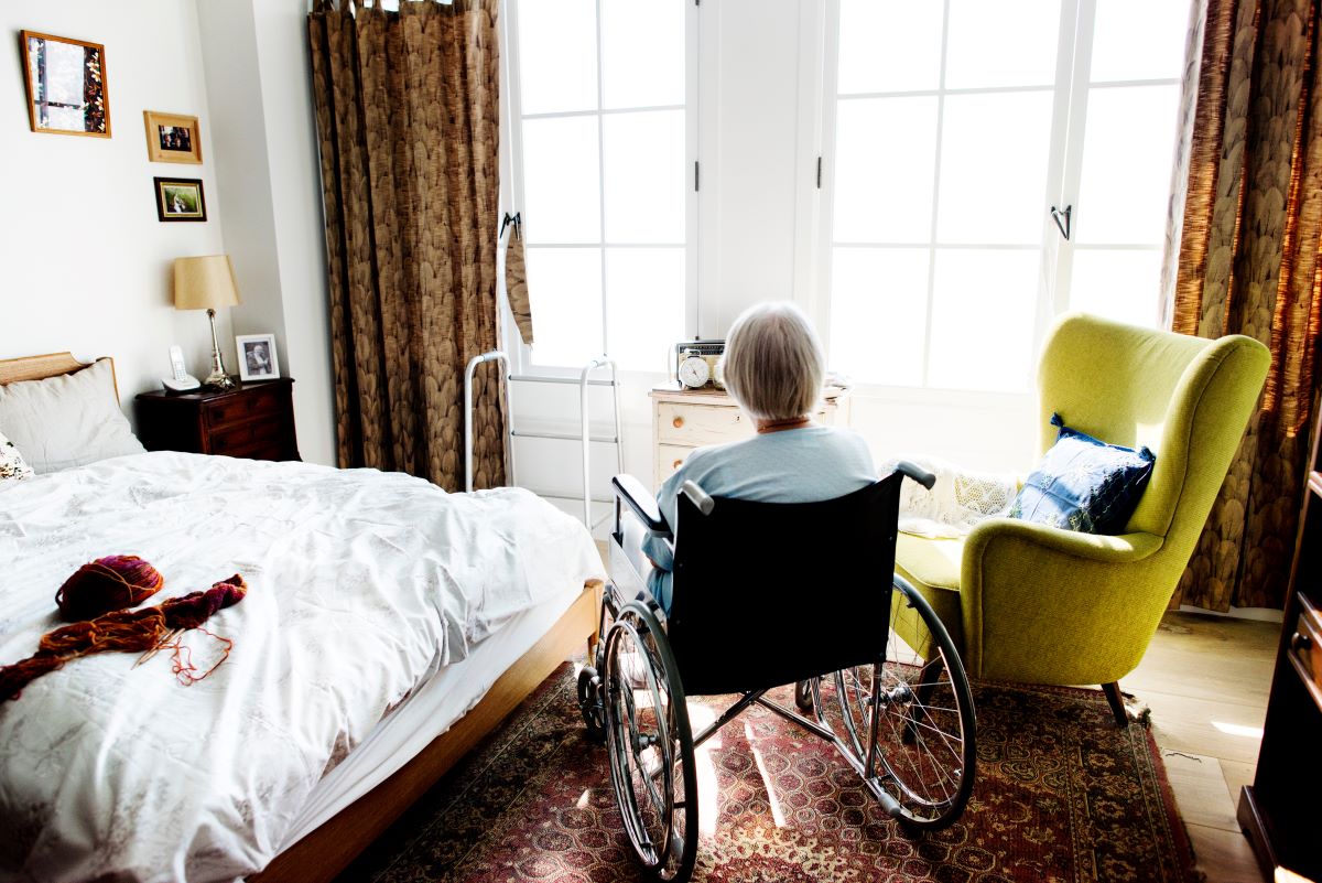 A nursing home resident sits in her wheelchair, looking out the window of her room.